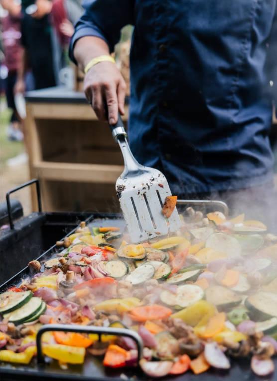 Nahaufnahme einer Grillplatte mit buntem Gemüse, das gerade mit einem Grillwender gewendet wird.