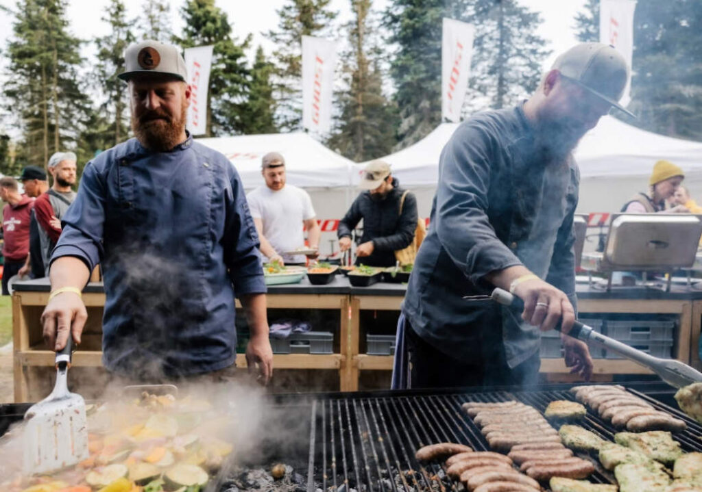 Zwei Männer in Kochkleidung grillen Gemüse und Würstchen auf einem großen Grill bei einer Outdoor-Veranstaltung.